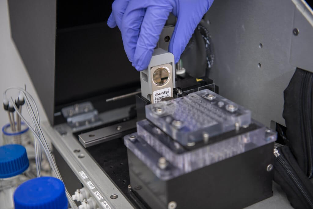 A close-up of a researcher wearing purple gloves handling a lateral flow diagnostic device in a laboratory, with scientific equipment and lab instruments visible in the background.