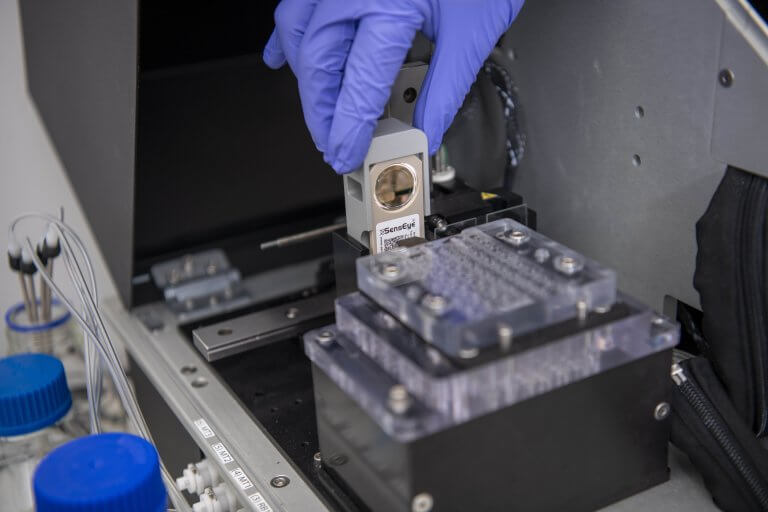 A close-up of a researcher wearing purple gloves handling a lateral flow diagnostic device in a laboratory, with scientific equipment and lab instruments visible in the background.